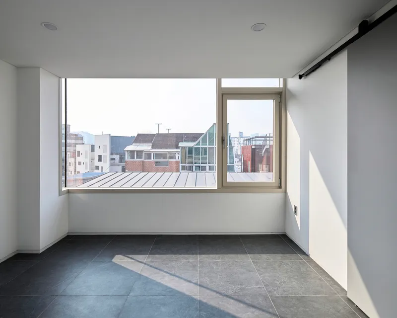 Empty room with large corner windows framing views of neighboring brick and glass buildings in daylight
