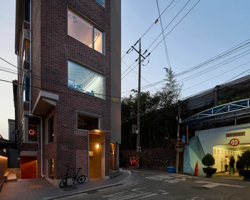 Red brick facade with stacked windows and entrance canopy beside a wooded hillside at twilight