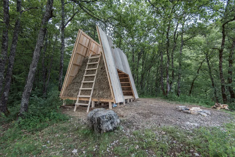 A-frame timber shelter with ladder and bundled straw infill standing in a forest clearing