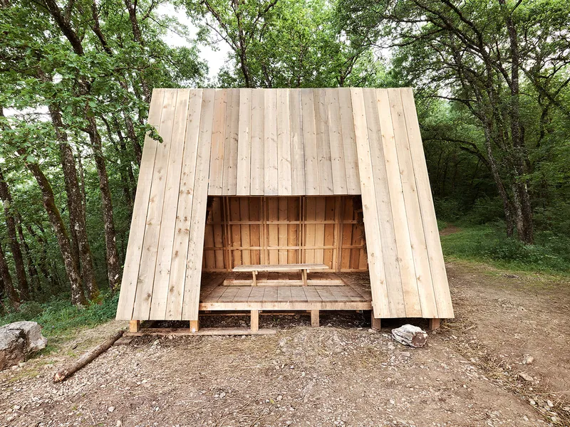 Front facade of the timber shelter showing the open entryway and bench inside the A-frame