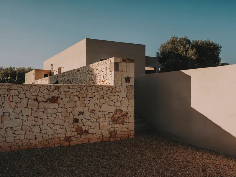 Layered stone and plaster volumes with a dry stone retaining wall beneath a clear sky