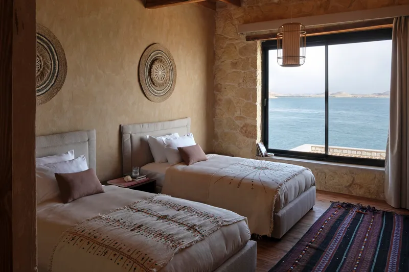 Twin beds with stone walls and exposed timber ceiling beams overlooking the water through large windows