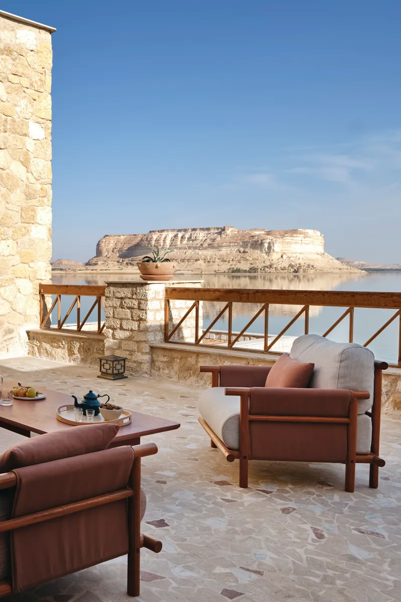 Limestone terrace with timber armchairs overlooking a distant mesa across calm water in bright daylight