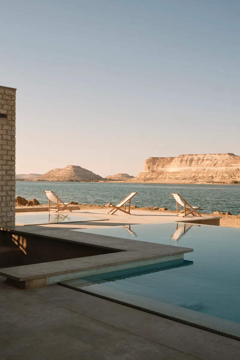 Infinity pool edge with three canvas deck chairs facing layered desert cliffs at golden hour