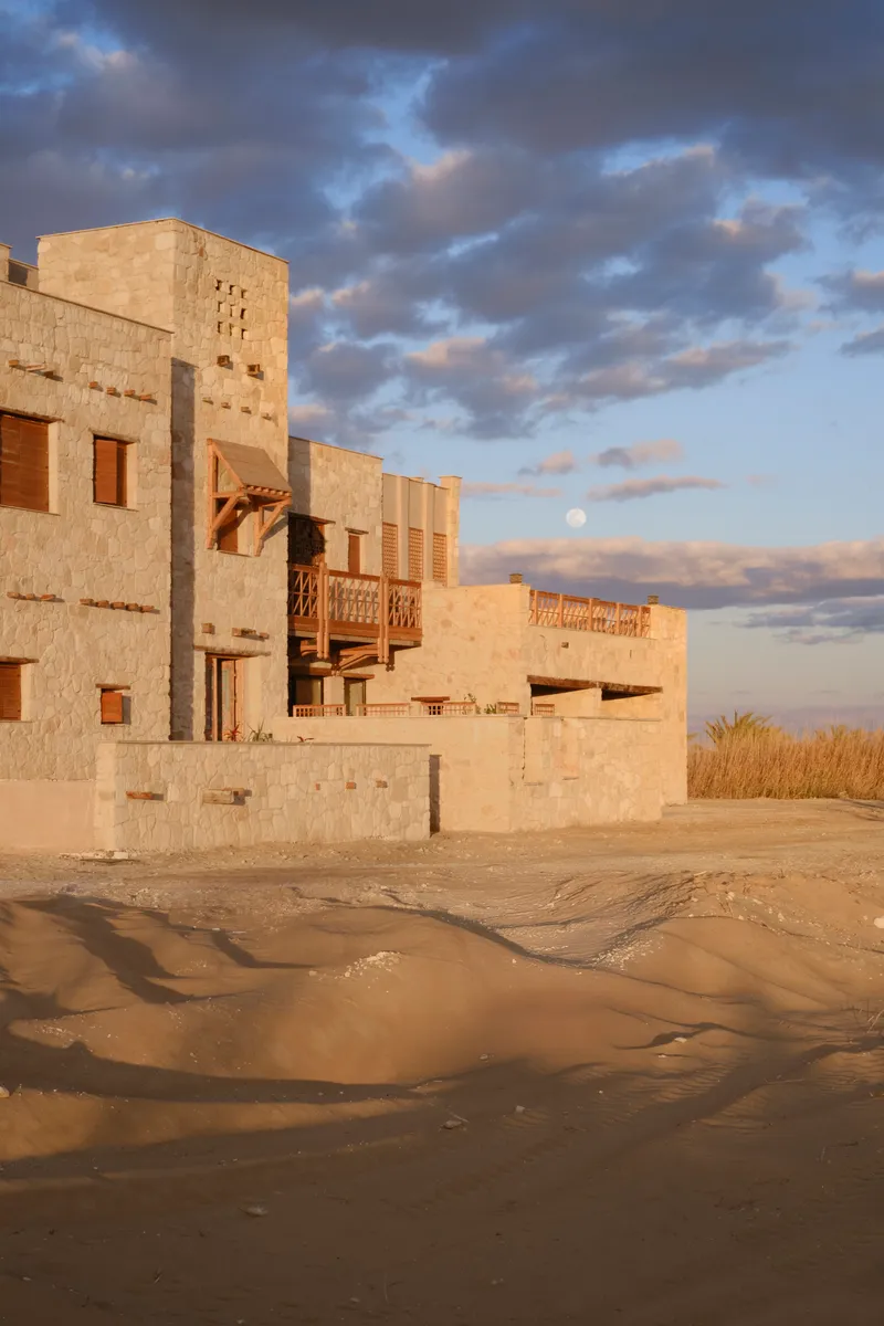 Clustered limestone volumes with timber balconies and openings rising from wet sand under evening clouds