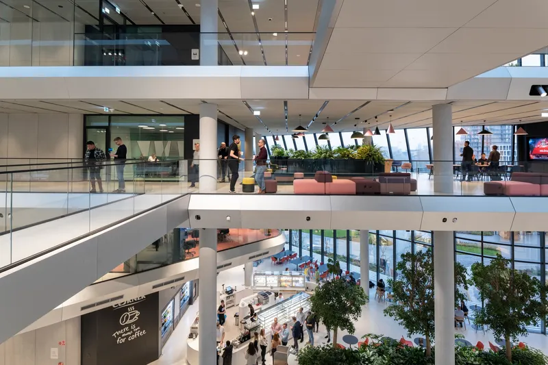 Multi-level interior atrium with white balconies, glass railings and people gathering around planted trees below