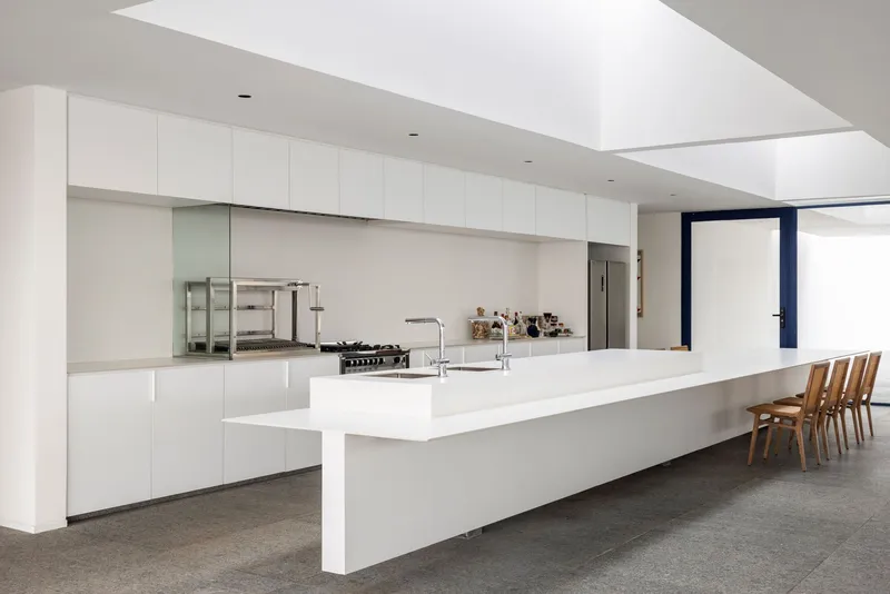 Long white kitchen bench with cantilevered dining extension under geometric ceiling volumes and natural daylight