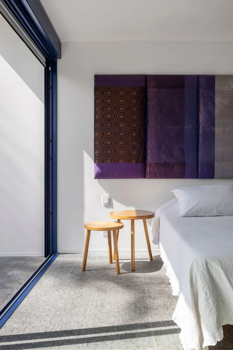 Bedroom corner with timber side tables and patterned textile artworks catching afternoon sunlight through dark-framed glazing