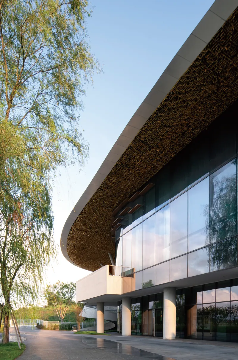 Glazed facade beneath a textured undulating roof canopy with white columns and willow trees at dusk