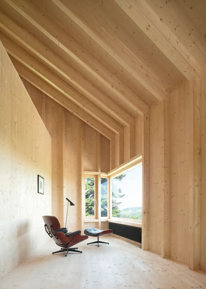 Interior view of exposed timber beam ceiling framing a corner window with lounge chair and floor lamp