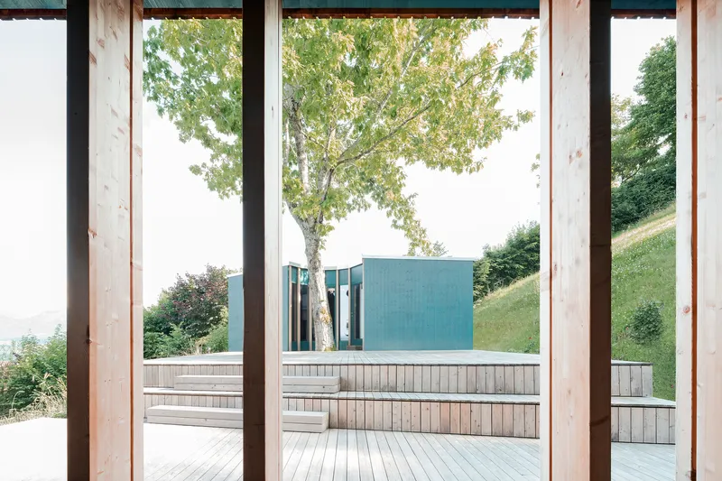 View from covered timber porch through black steel columns toward terraced deck and blue metal volume