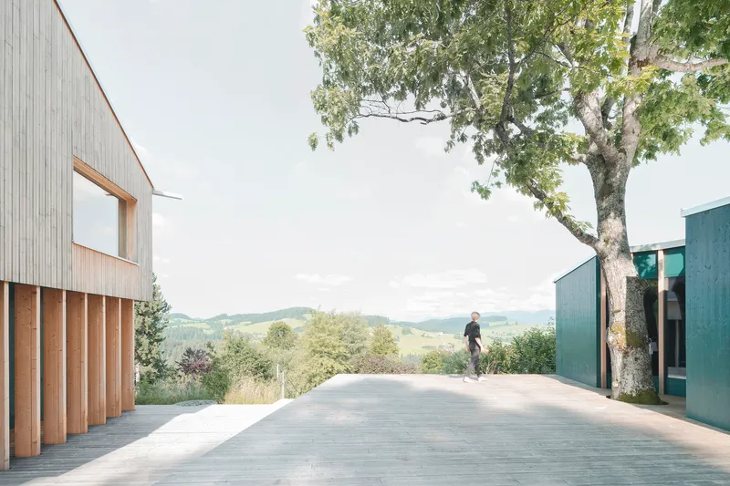Concrete terrace between vertical timber-clad volume and blue metal pavilion with two figures beneath a tree