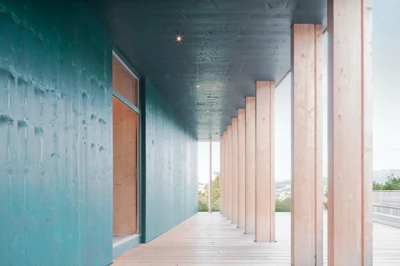 Covered colonnade with timber columns between blue stained wood walls and decking under grey sky