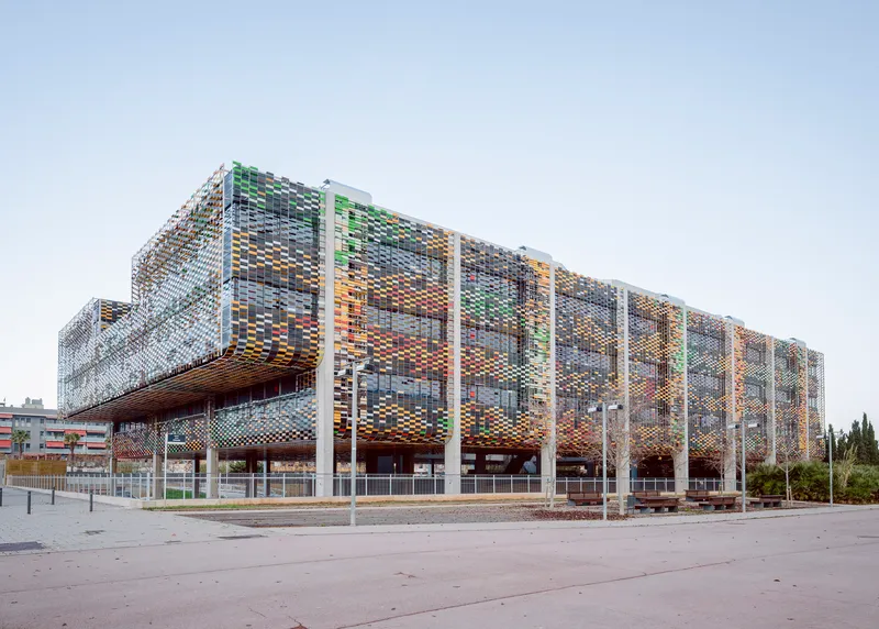 Corner view of the multi-coloured perforated metal screen wrapping the elevated volume above a paved plaza