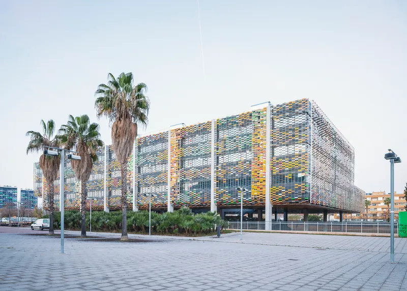 Perforated facade with multicoloured ceramic panels on white columns viewed from a paved plaza with palm trees
