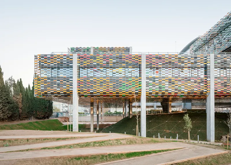 Elevated structure with multicoloured ceramic screen cladding supported by white concrete columns above a landscaped slope