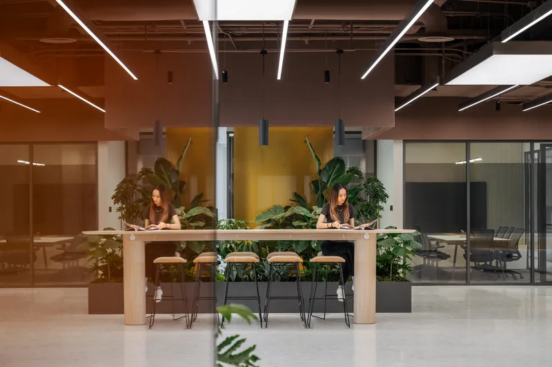 Shared work table with light wood top surrounded by planters and bar stools under linear ceiling lights