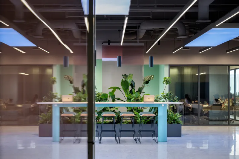 Open office lounge with a high-top table, wire-frame stools and large tropical plants beneath exposed ceiling