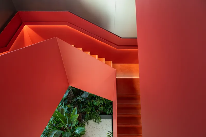 Orange-lit stairwell with folded red walls descending past a ground-level planter bed with ferns and tropical plants