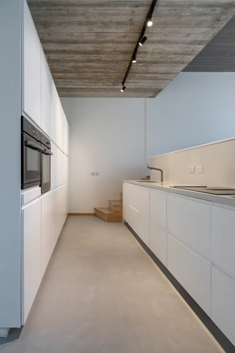 Galley kitchen with white cabinetry and timber steps beneath board-formed concrete ceiling and black track lighting