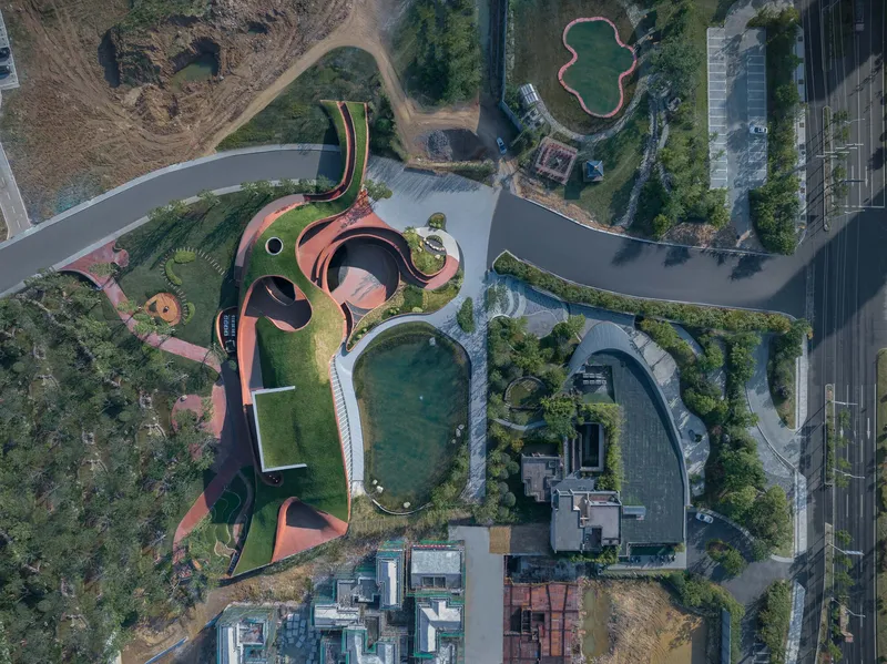 Aerial view of the curving rust-colored roof with circular voids and planted green roof among water features and courtyards