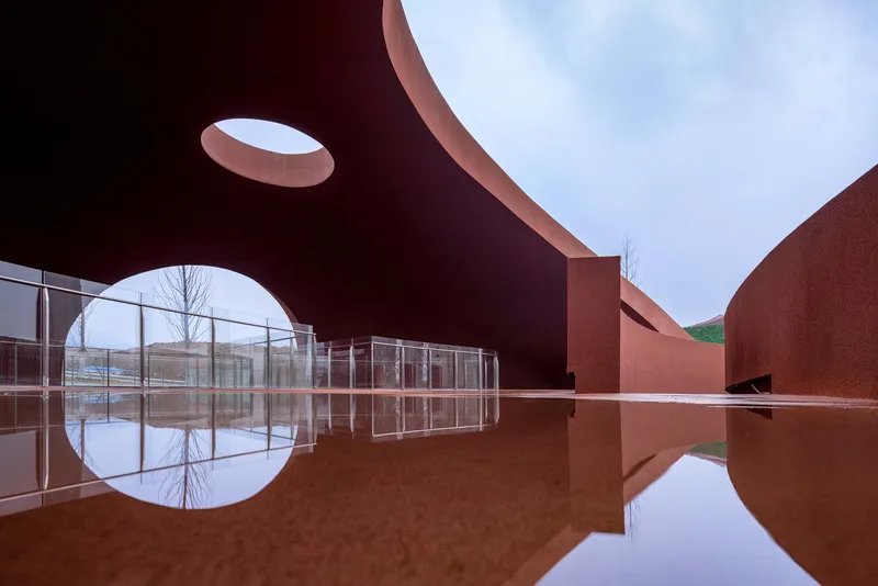 Reflecting pool mirroring the curved red canopy with circular openings and glazed gallery beyond