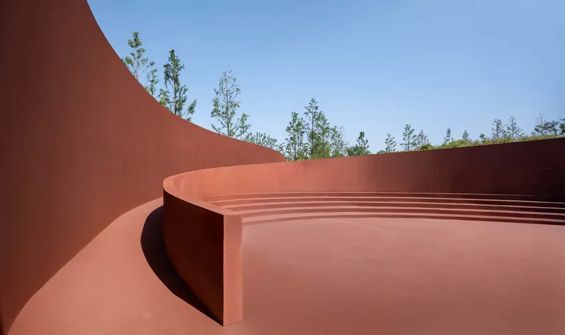 Terraced amphitheater steps within curved red walls open to planted trees and sky above