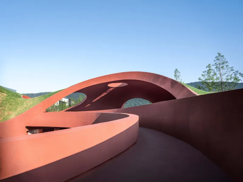 Rust-colored roof terrace with circular and oval openings framing views of hillside vegetation under clear blue sky
