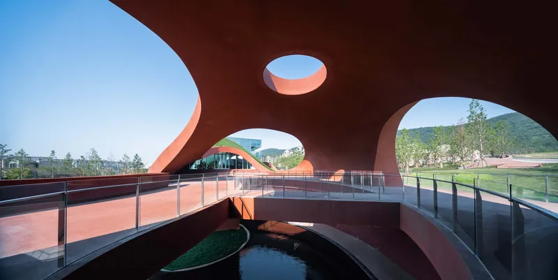 View through circular and oval roof openings toward glass pavilion and landscape beyond from beneath rust-colored canopy