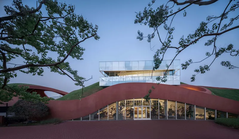 Main entrance with glazed facade beneath the sweeping roof canopy framed by mature trees at dusk
