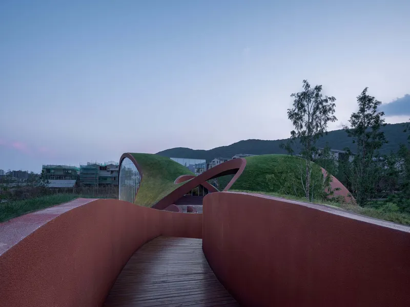 Timber-decked ramp between terracotta walls leading toward grass-covered roof forms at twilight