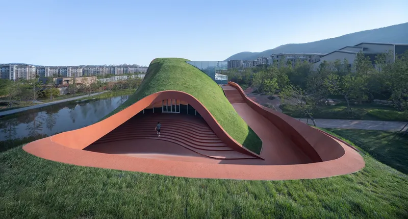 Sunken amphitheater with timber seating beneath a grass-covered roof beside a reflecting pond