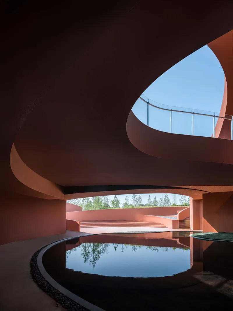 Circular reflecting pool beneath flowing red concrete shell with triangular skylight openings above