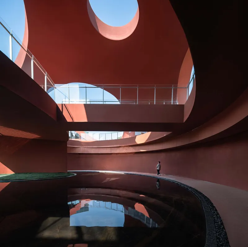 Curving water basin with person walking along edge beneath layered red concrete walls and circular skylight