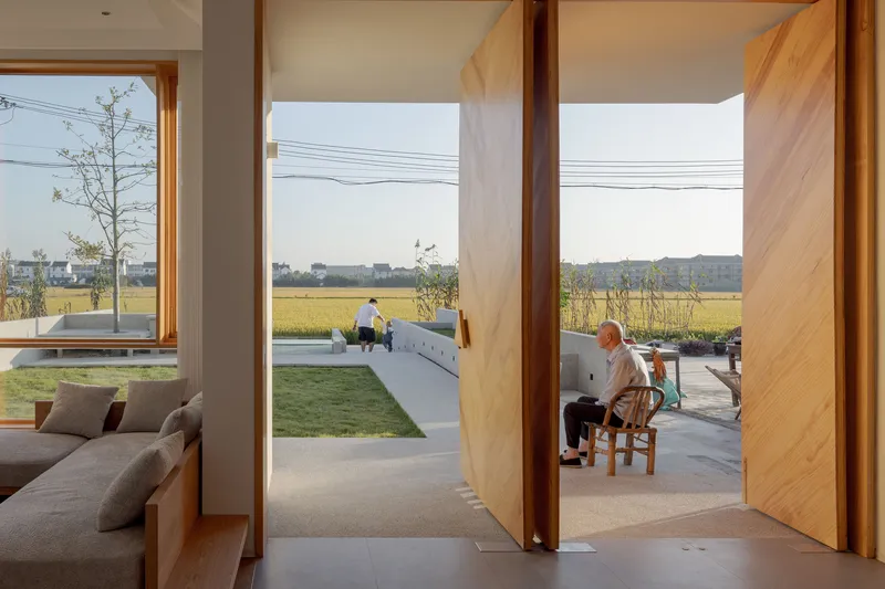 Interior view through pivoting timber doors to a terrace where residents sit overlooking the rice fields