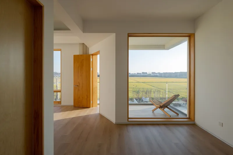 Interior bedroom with timber framed window framing a folding chair on the terrace and distant agricultural fields