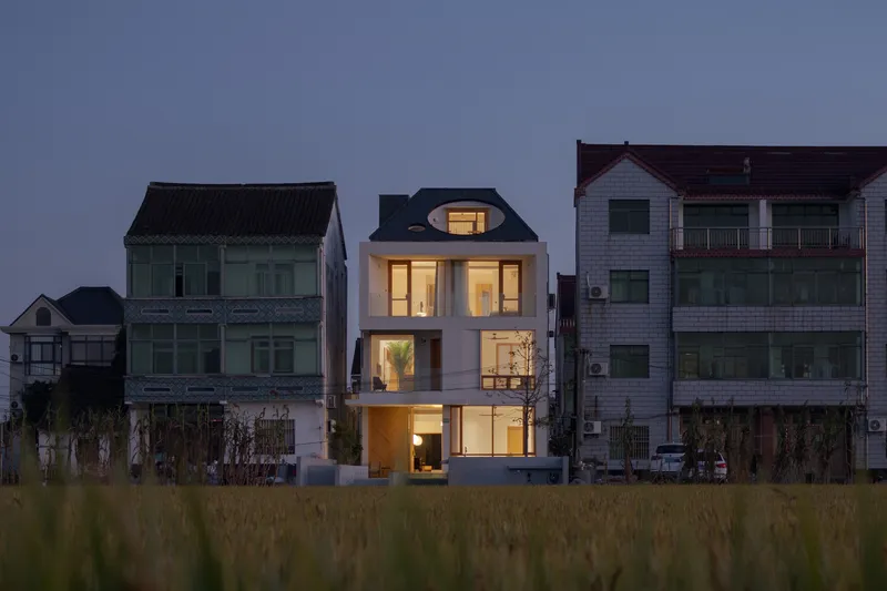 White rendered facade with illuminated arched dormer window nestled among neighboring houses at dusk