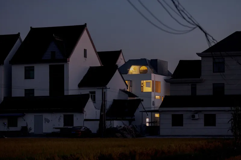 Street view of the glowing arched dormer rising above silhouetted rooflines under overhead power lines