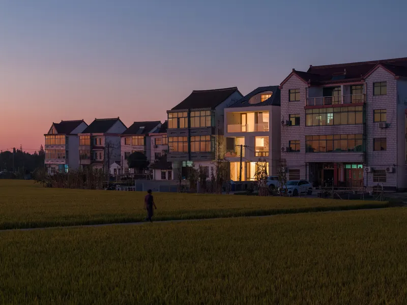 Figure walking through rice paddies at sunset with illuminated house facade visible beyond green field