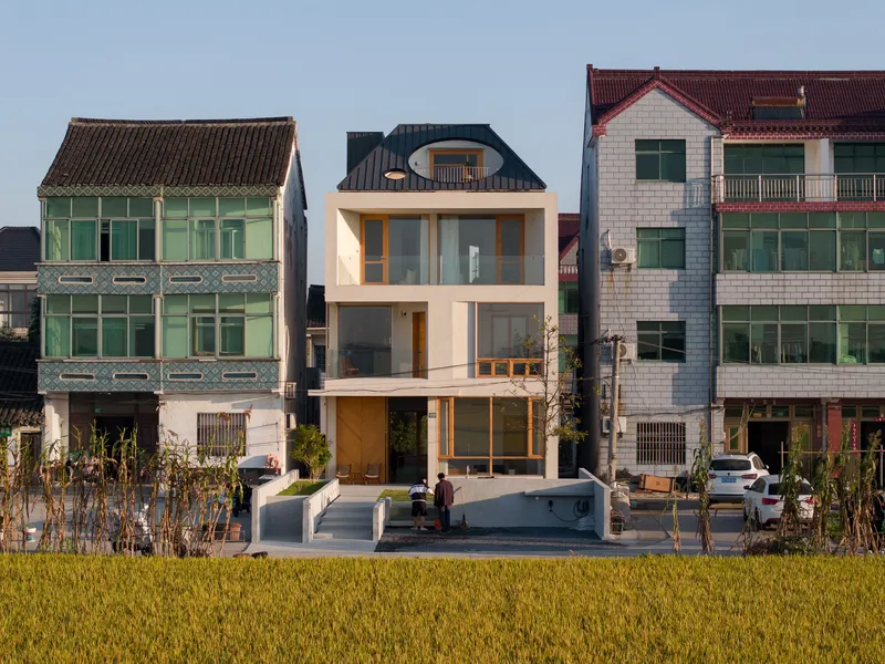 Street facade with white render and timber framed openings standing among older houses and a golden rice paddy