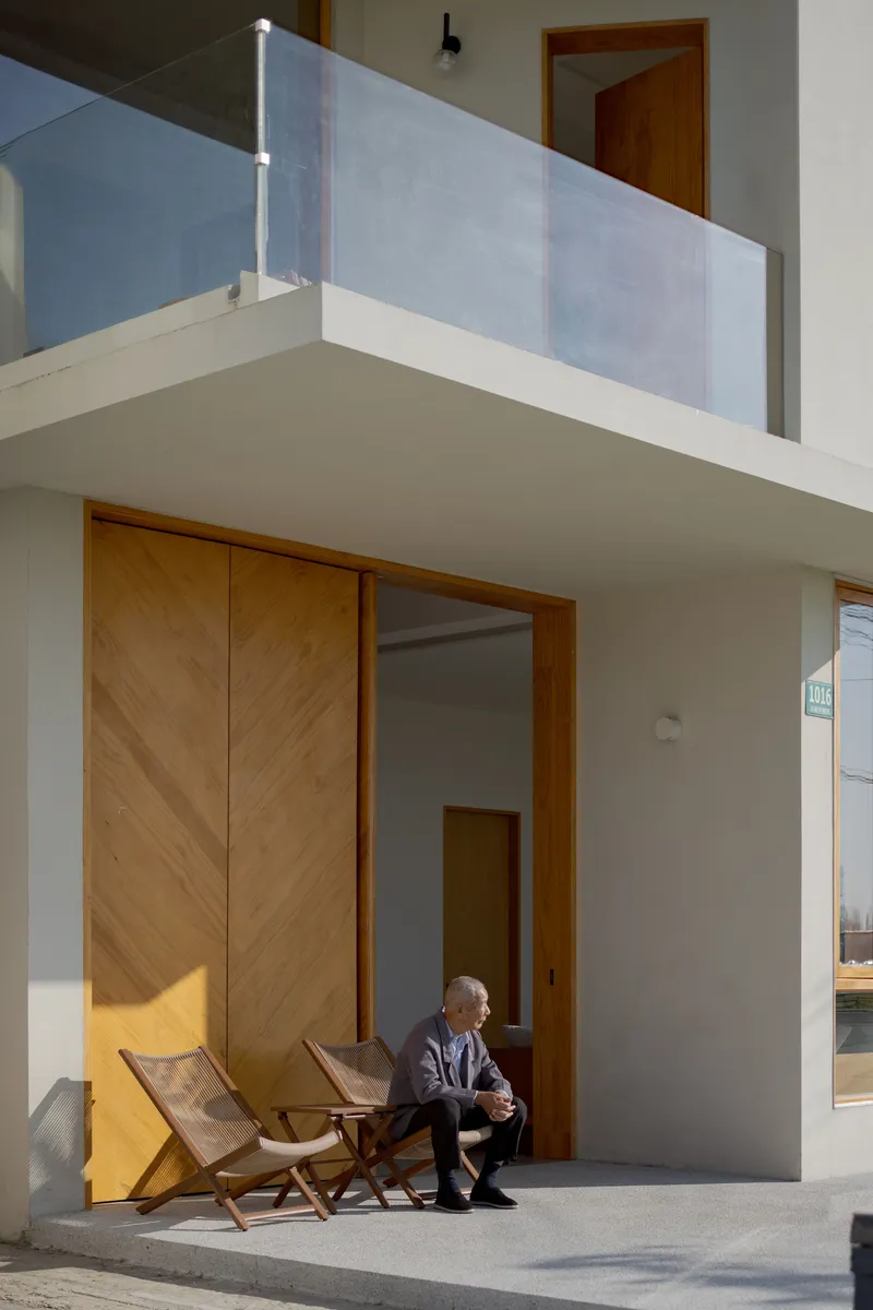 Covered terrace with diagonal timber doors and a seated figure beside folding chairs in afternoon sunlight