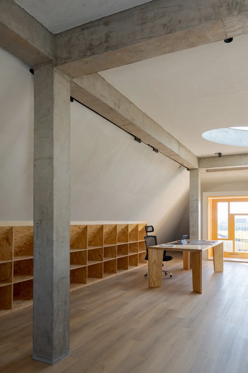 Interior workspace with exposed concrete beam spanning above built-in plywood shelving and timber desk