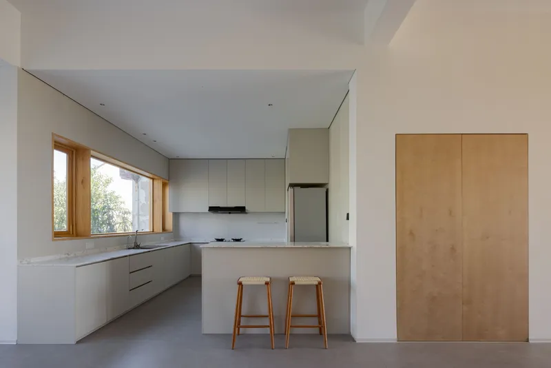 Minimalist kitchen with white island, plywood cabinetry and timber-framed window overlooking greenery