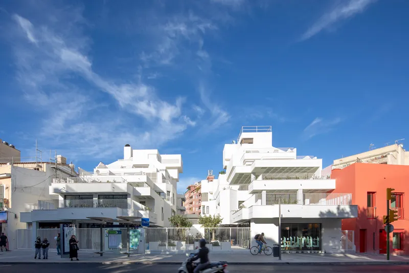 Street view of white stepped housing volumes with vertical louvers and pedestrians below wispy clouds