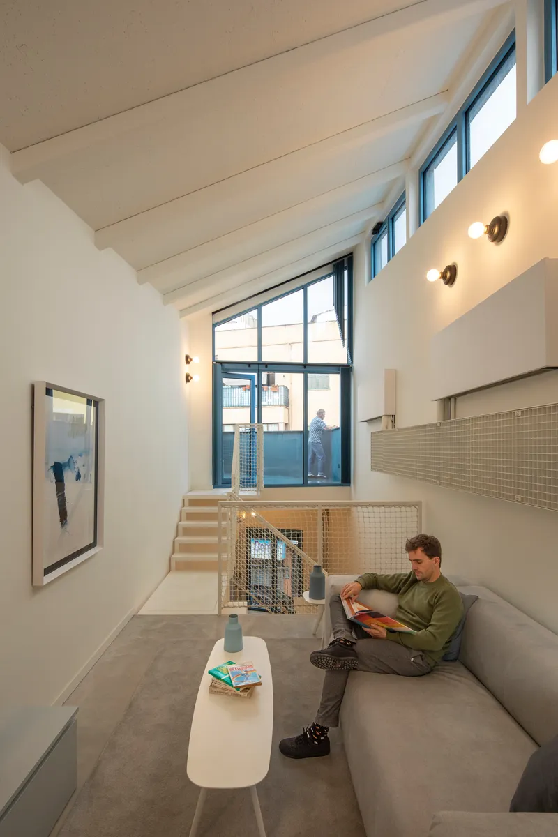 Double-height living room with painted timber ceiling beams and person reading on sofa below mezzanine