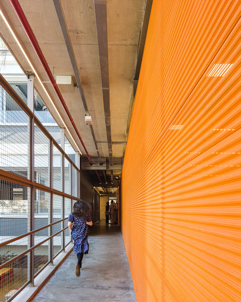 Corridor with exposed concrete ceiling, orange corrugated wall panels and mesh balustrade overlooking the courtyard