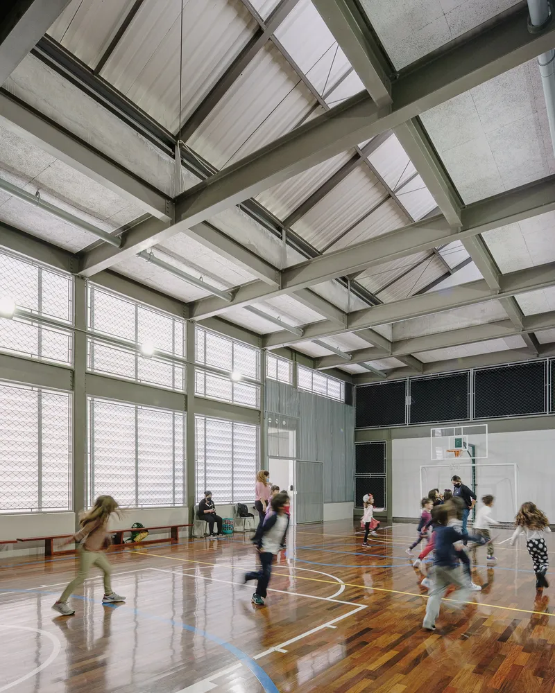 Interior gymnasium with timber floor, exposed concrete ceiling trusses and children playing beneath wire mesh windows