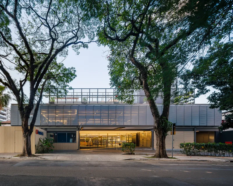 Street facade with translucent upper level, timber cladding at entry, and mature trees flanking the sidewalk at dusk
