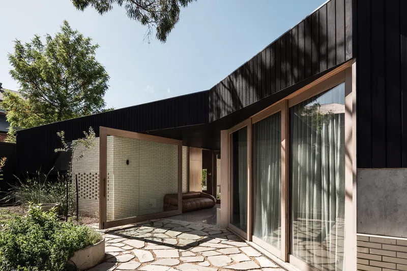 Courtyard with broken flagstone paving, cream brick screening wall and timber-framed glazing under a black timber soffit
