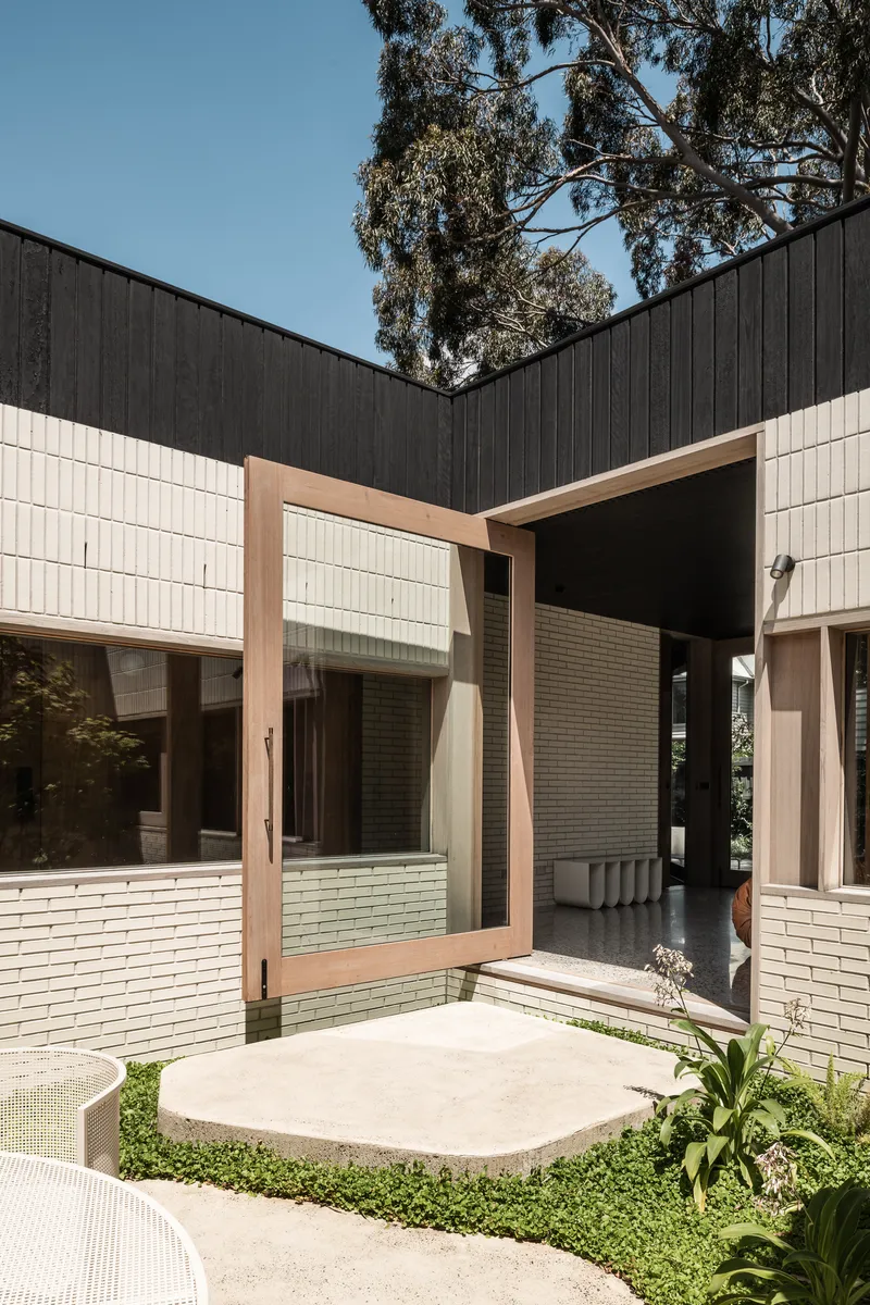 Curved concrete paving inset with groundcover leading to timber-framed opening in cream brick volume beneath dark cladding